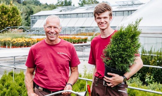 2 Gärtner in rotem Oberteil stehen in einem Garten, einer hält einen Grünen Baum, der andere einen Schlauch in der Hand.