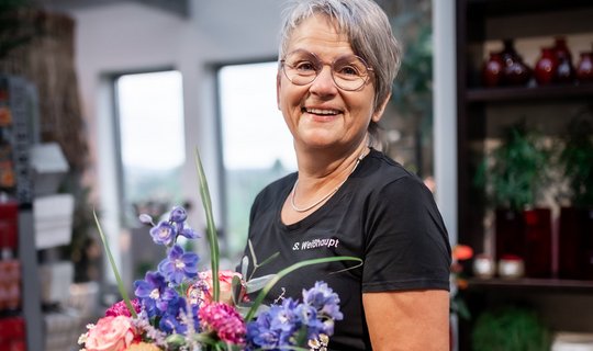 Floristinmeisterin steht mit blau, rosa orangenem Blumenstrauß im Bild.
