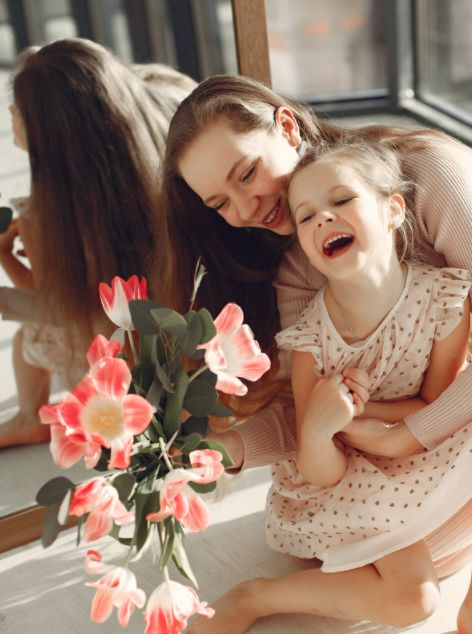 Lachende Mutter und Tochter mit Strauß rosa Tulpen in heller Umgebung, beide wirken fröhlich und vertraut.
